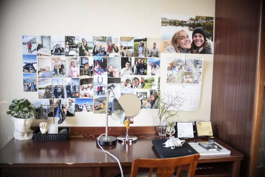 Skrivebord med bilder av venner og familie hengt opp på veggen, en speil, bordlampe, planter og kalendere.