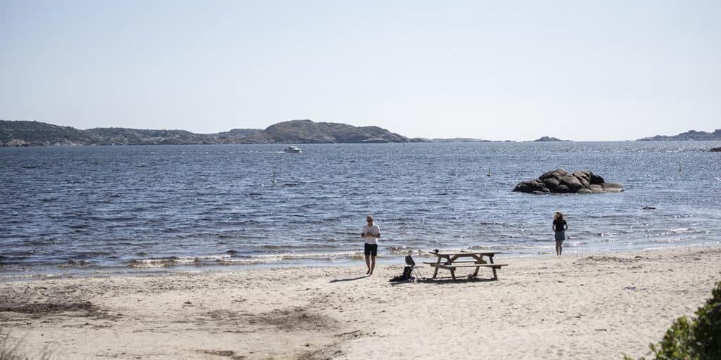 Strand med to personer ved vannkanten, et piknikbord og øyer i horisonten under en klar himmel.
