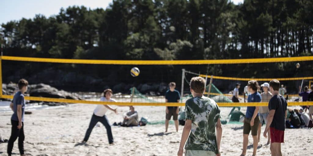 Unge som spiller volleyball på en sandstrand med skog i bakgrunnen.