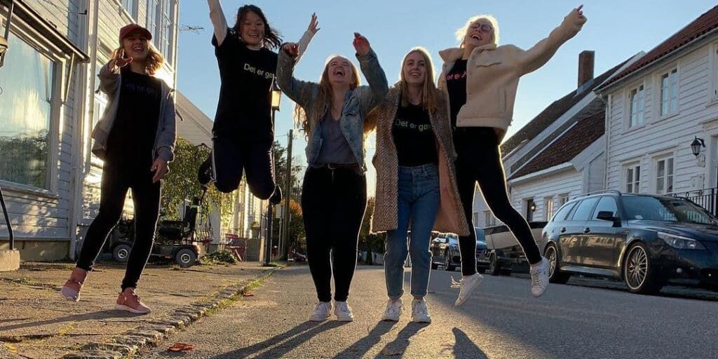 Fem unge kvinner hopper og smiler i en solrik gate med hvite hus og parkerte biler.