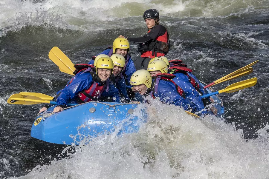 Seks personer iført gule hjelmer og blå redningsvester i en blå raftbåt navigerer gjennom fosser og bølger i strykende elv.