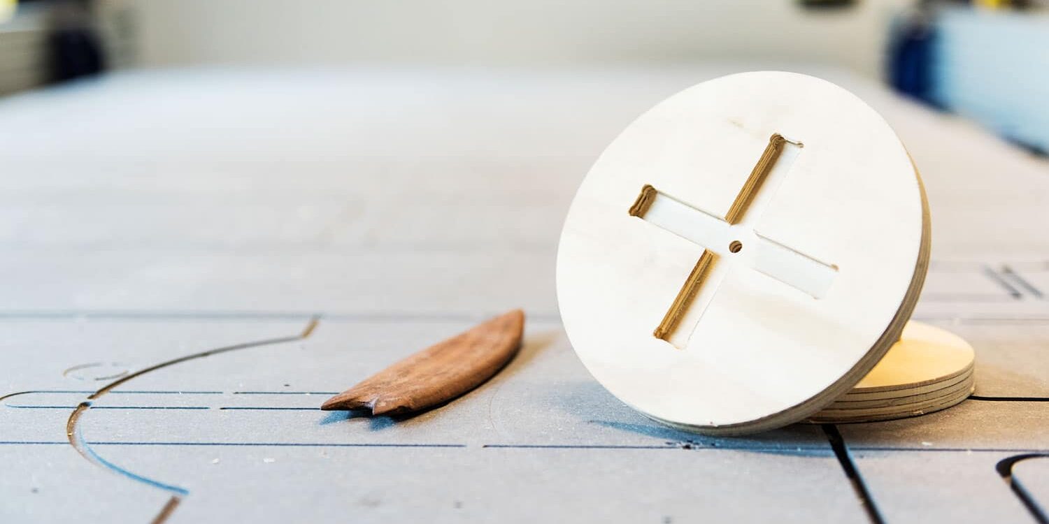 En rund, lys trefiksingsplate med kryssformet utskjæring, ved siden av et lite, buet treverkøy, liggende på en trefresingsmaskin.