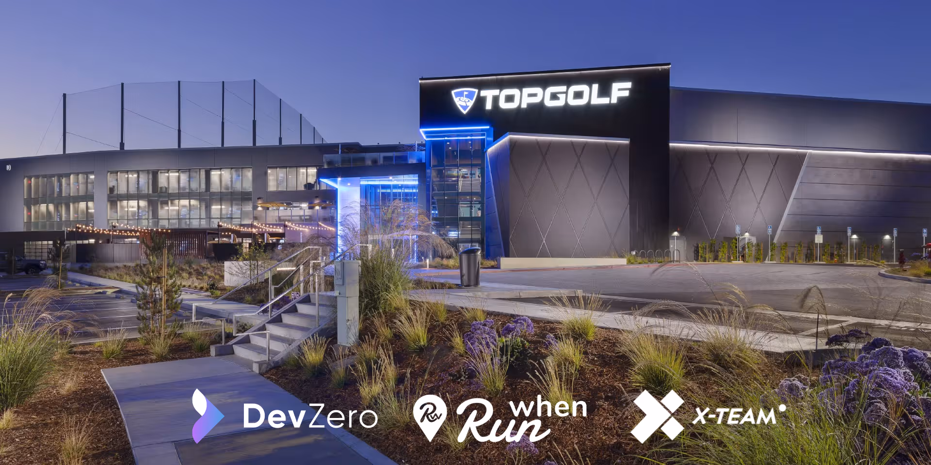 Exterior view of a Topgolf building illuminated at dusk with landscaped greenery in the foreground.