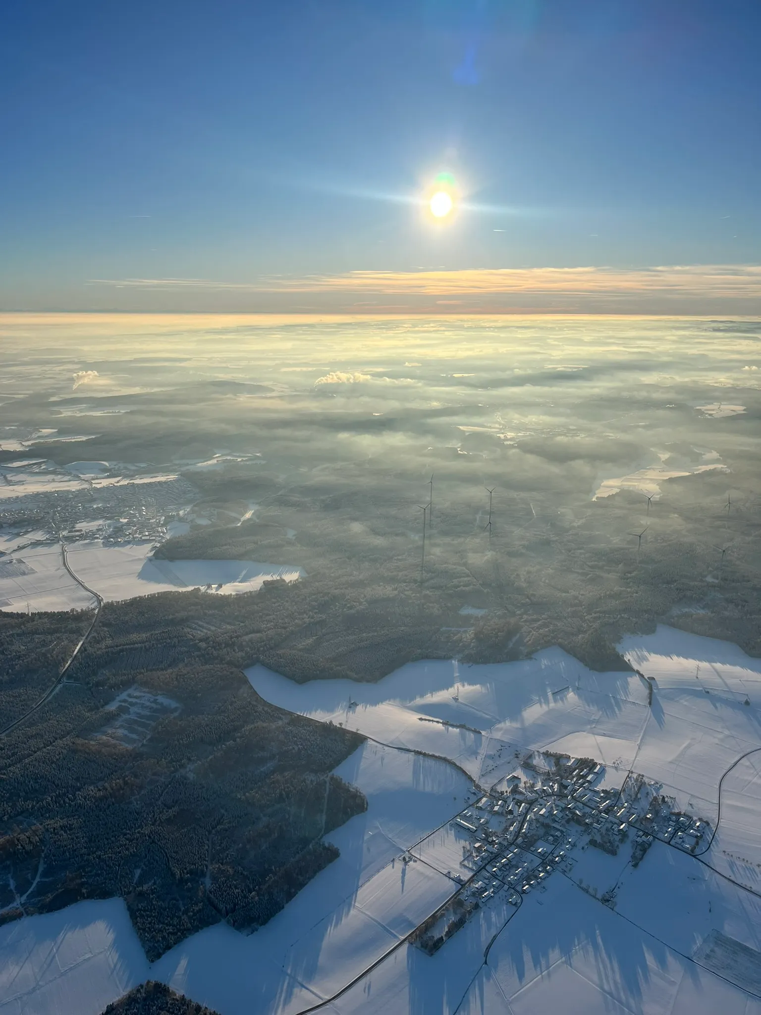 Luftaufnahme einer verschneiten Landschaft mit Wald, Windrädern und Sonne über einem Nebelmeer am klaren Himmel.