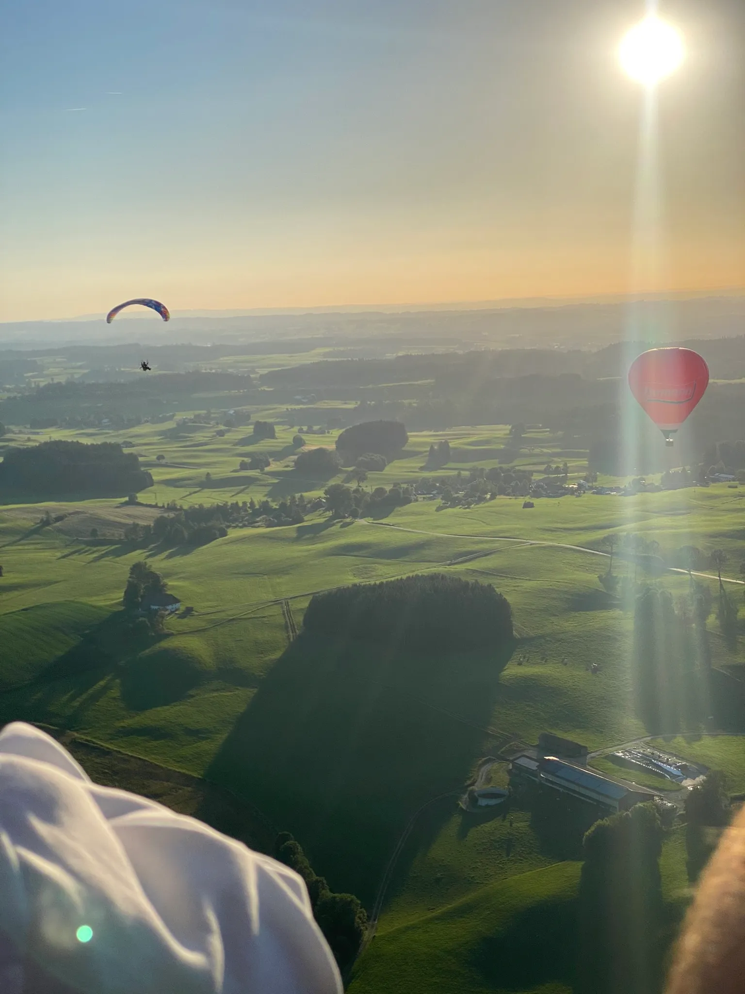 Blick aus der Luft auf grüne Felder bei Sonnenuntergang mit einem Gleitschirm und einem roten Heißluftballon am Himmel.