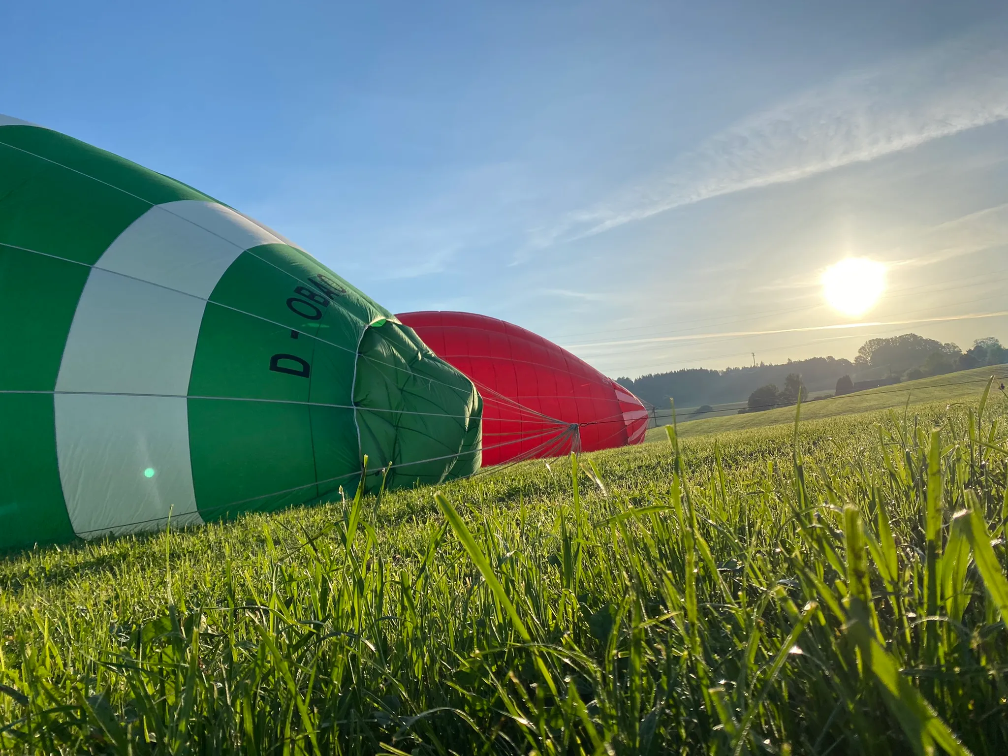 Zwei Heißluftballons, grün-weiß und rot, liegen auf einem grasbewachsenen Feld bei Sonnenaufgang.