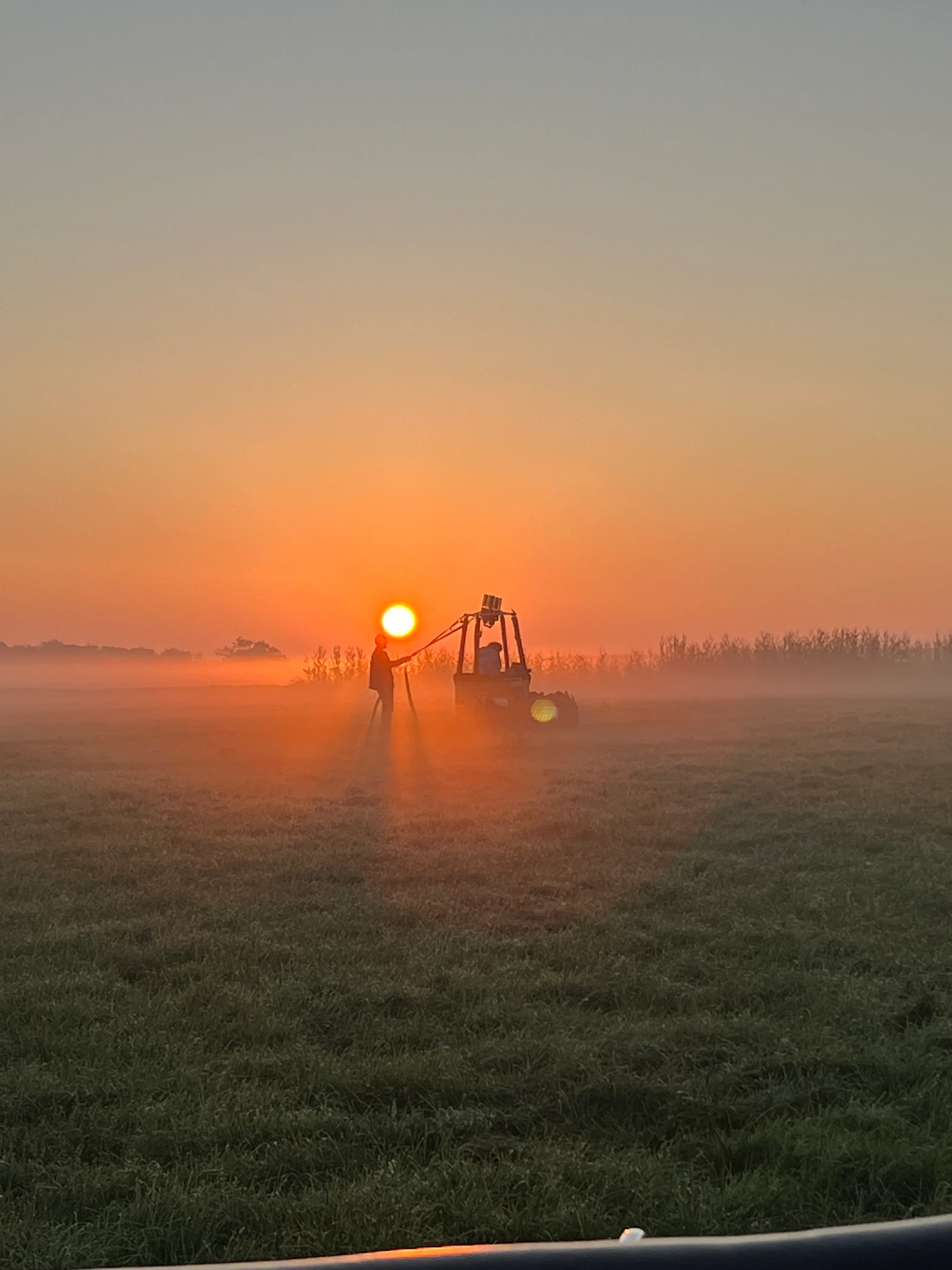 Silhouette eines Mannes neben landwirtschaftlicher Maschine auf nebeligem Feld bei Sonnenaufgang.