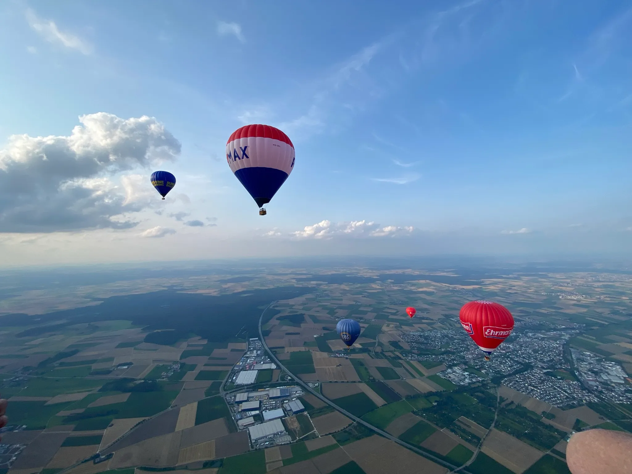 Heißluftballons mit unterschiedlichen Farben und Logos schweben über einem grünen und braunen Patchwork-Landschaft unter blauem Himmel mit Wolken.