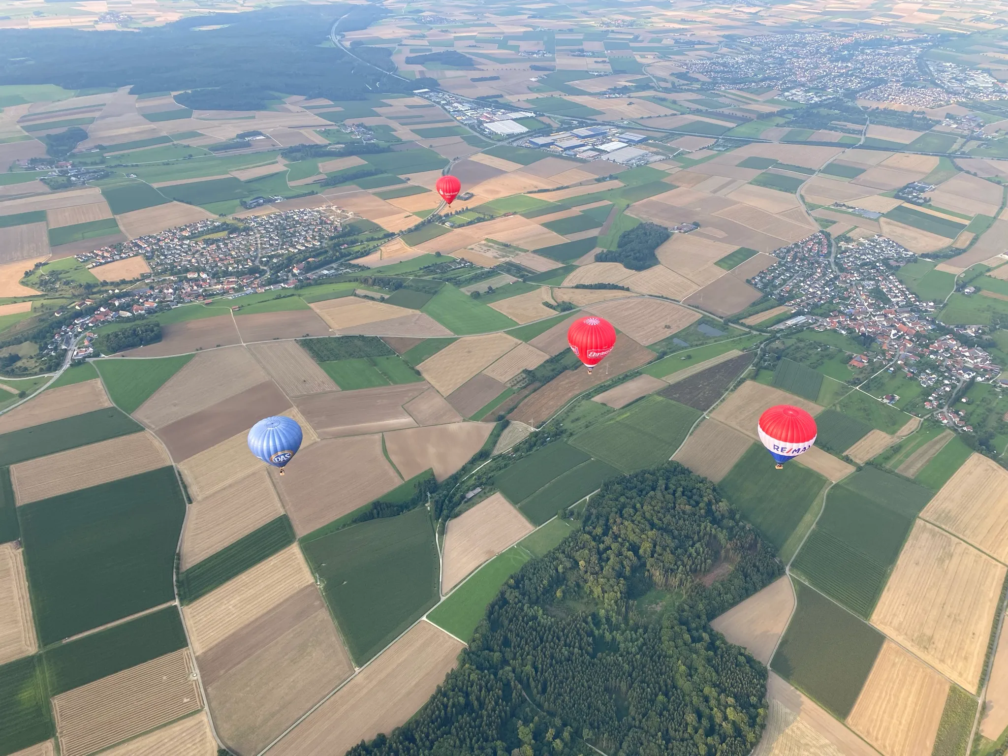 Luftaufnahme von Feldern und Dörfern mit vier Heißluftballons in verschiedenen Farben über der Landschaft.