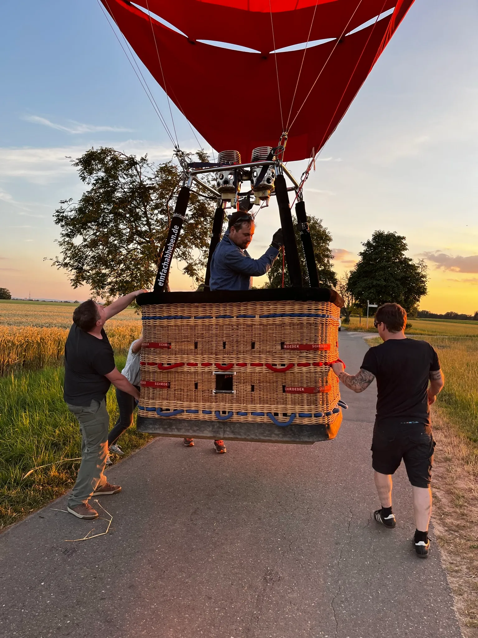 Drei Personen schieben einen mit einem roten Heißluftballon verbundenen Weidenkorb auf einer Landstraße bei Sonnenuntergang.