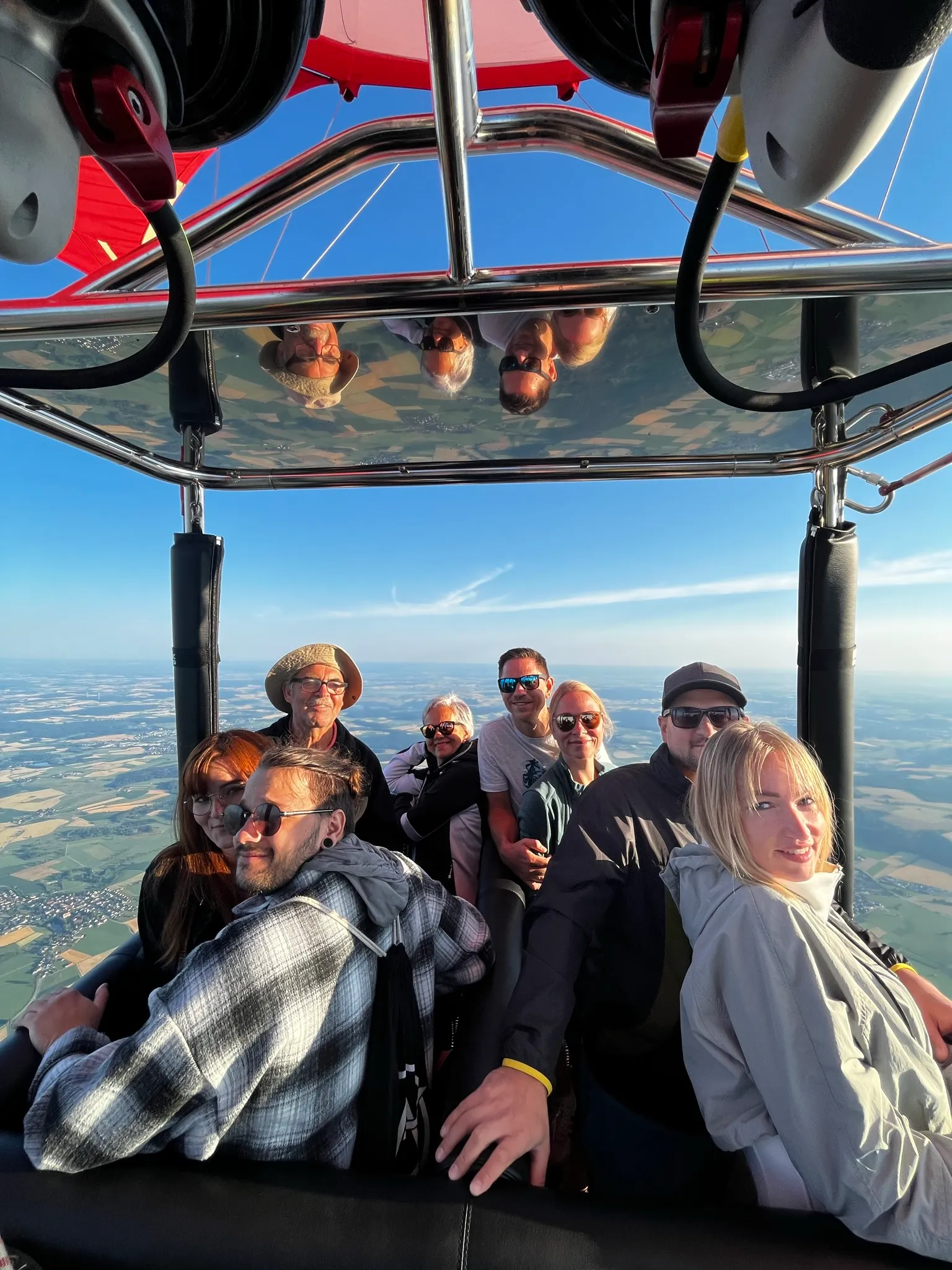 Acht Personen in einem Heißluftballonkorb, die bei klarem Himmel und Landschaft im Hintergrund lächeln.