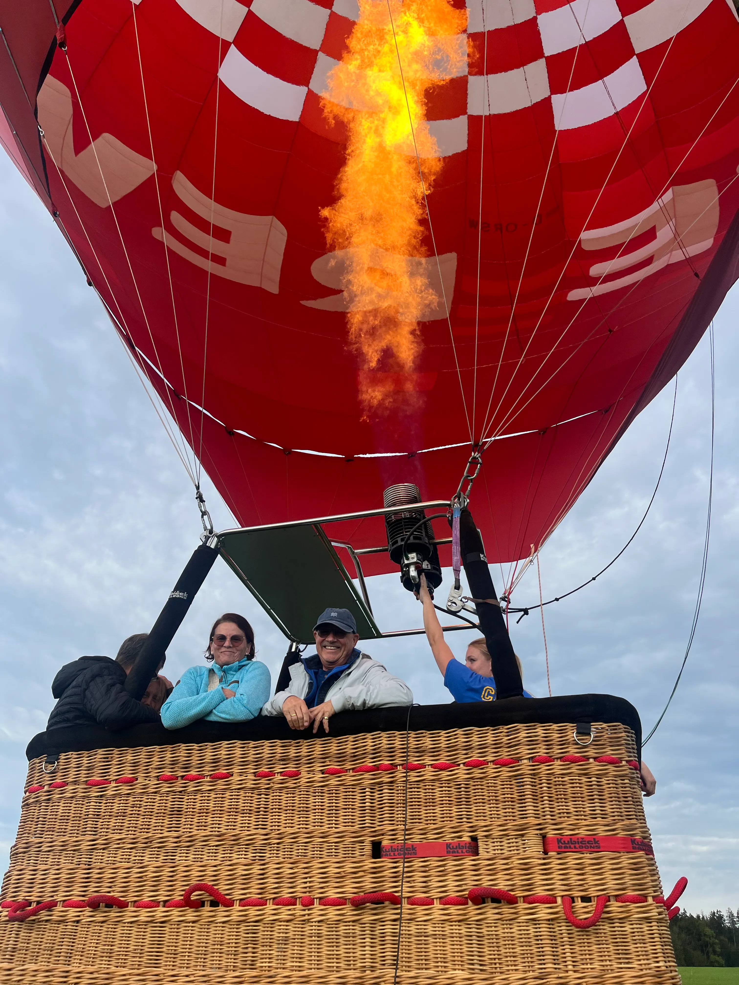 Ein Heißluftballon mit rotem Muster und Flamme, während mehrere Personen im Korb stehen und lächeln.