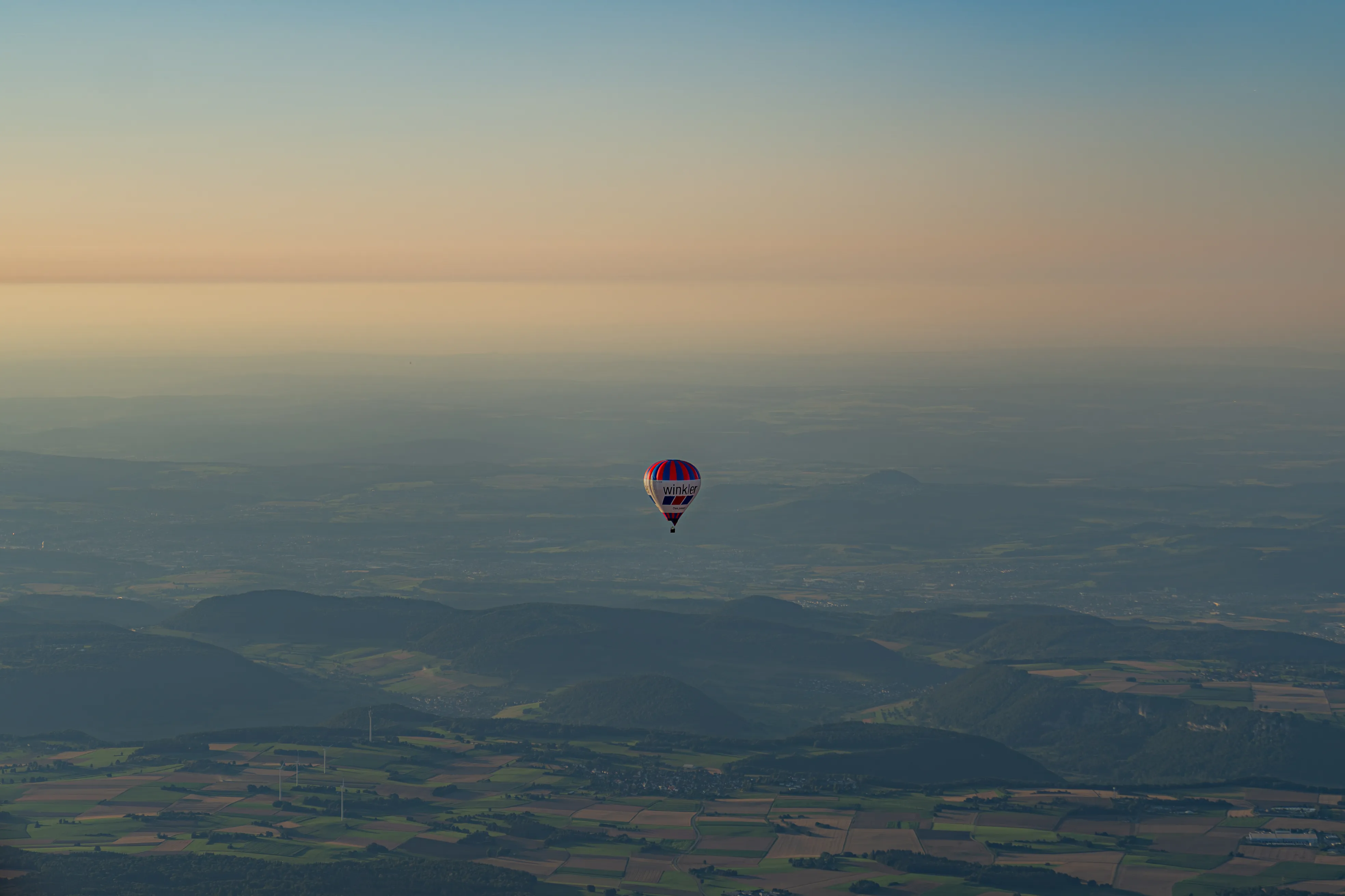 Heißluftballon mit der Aufschrift 'winkler' schwebt über einem weiten Tal mit Feldern und bewaldeten Hügeln im Dämmerlicht.