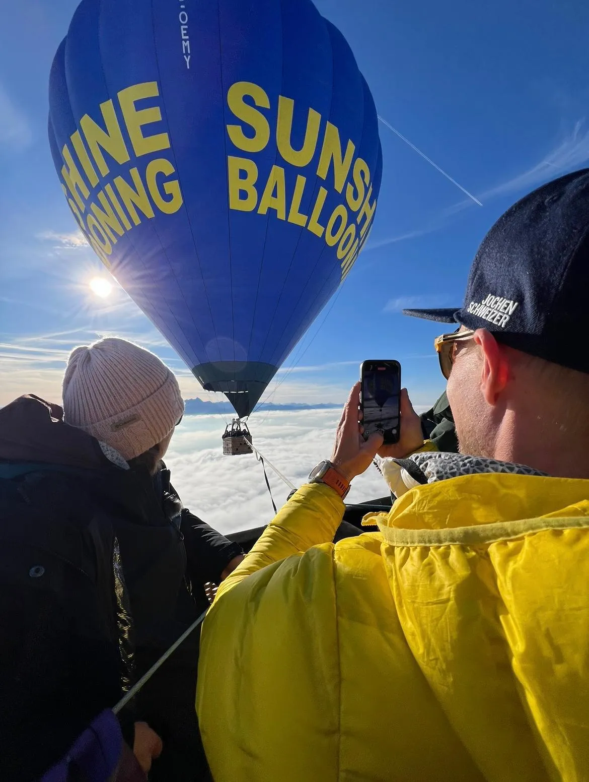 Zwei Personen in einem Heißluftballonkorb nehmen ein Foto von einem blauen Heißluftballon mit der Aufschrift 'SUNSHINE BALLOONING' über einer Wolkendecke bei sonnigem Himmel auf.
