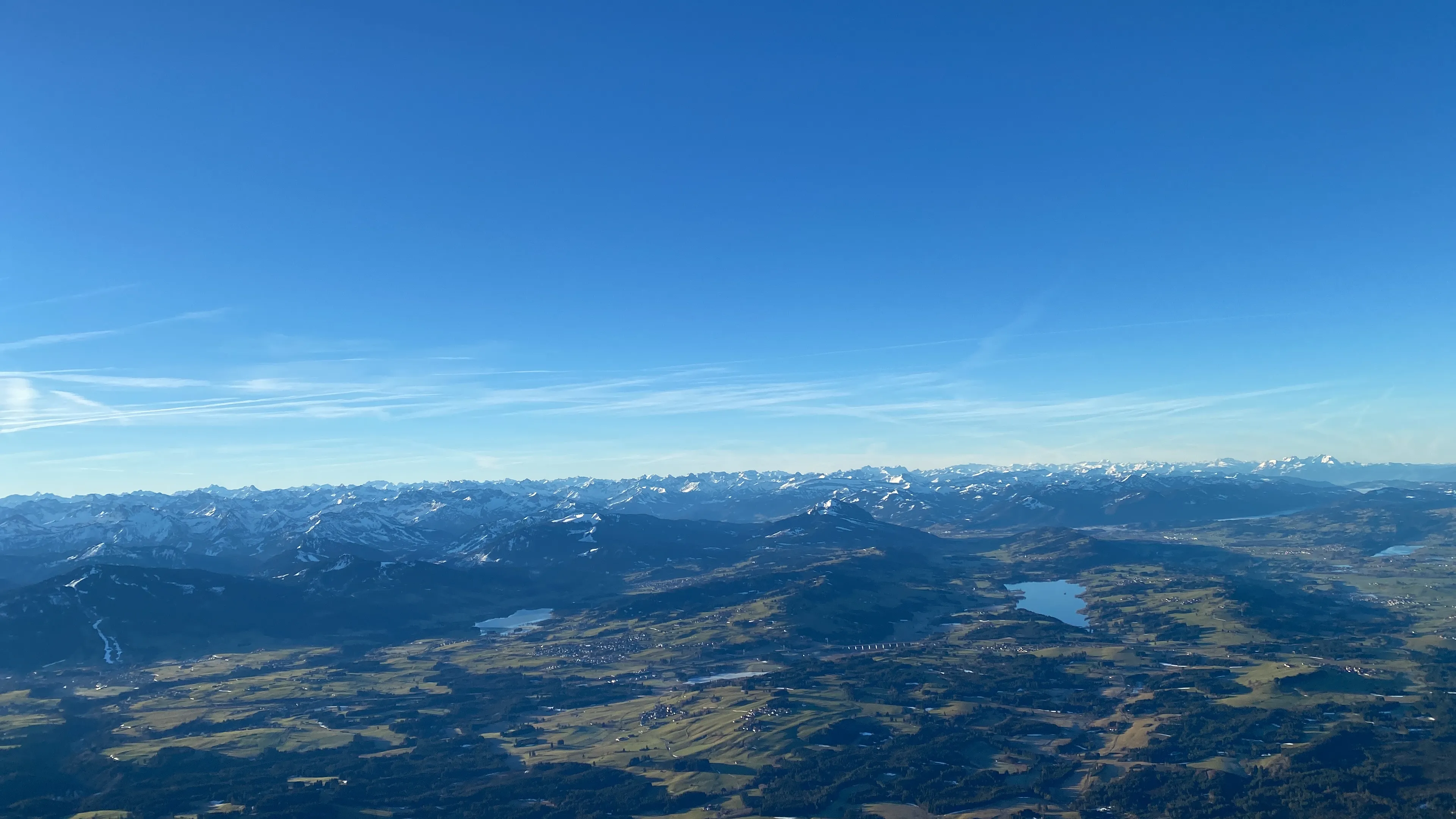 Blick auf eine grüne Hügellandschaft mit Seen vor einer langen Bergkette unter klarem blauem Himmel.