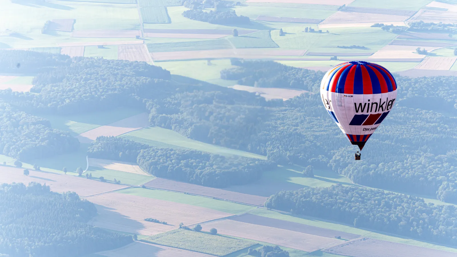 Ein Heißluftballon mit rot-blau-weißen Streifen und der Aufschrift „winkler“ über landwirtschaftlichen Feldern und Waldgebieten.