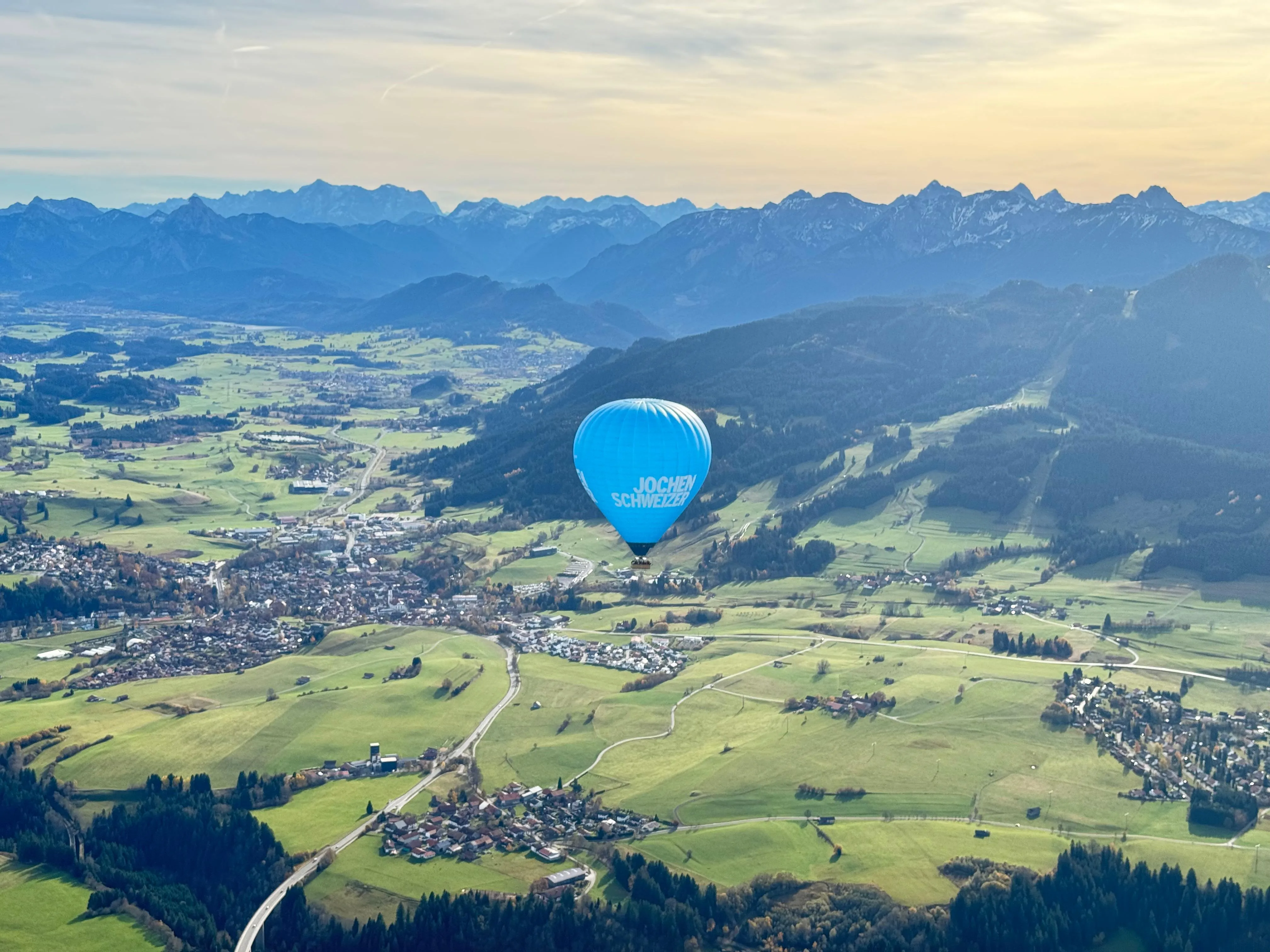 Blauer Heißluftballon mit der Aufschrift 'Jochen Schweizer' über grünem Tal vor Alpenpanorama bei Sonnenuntergang.