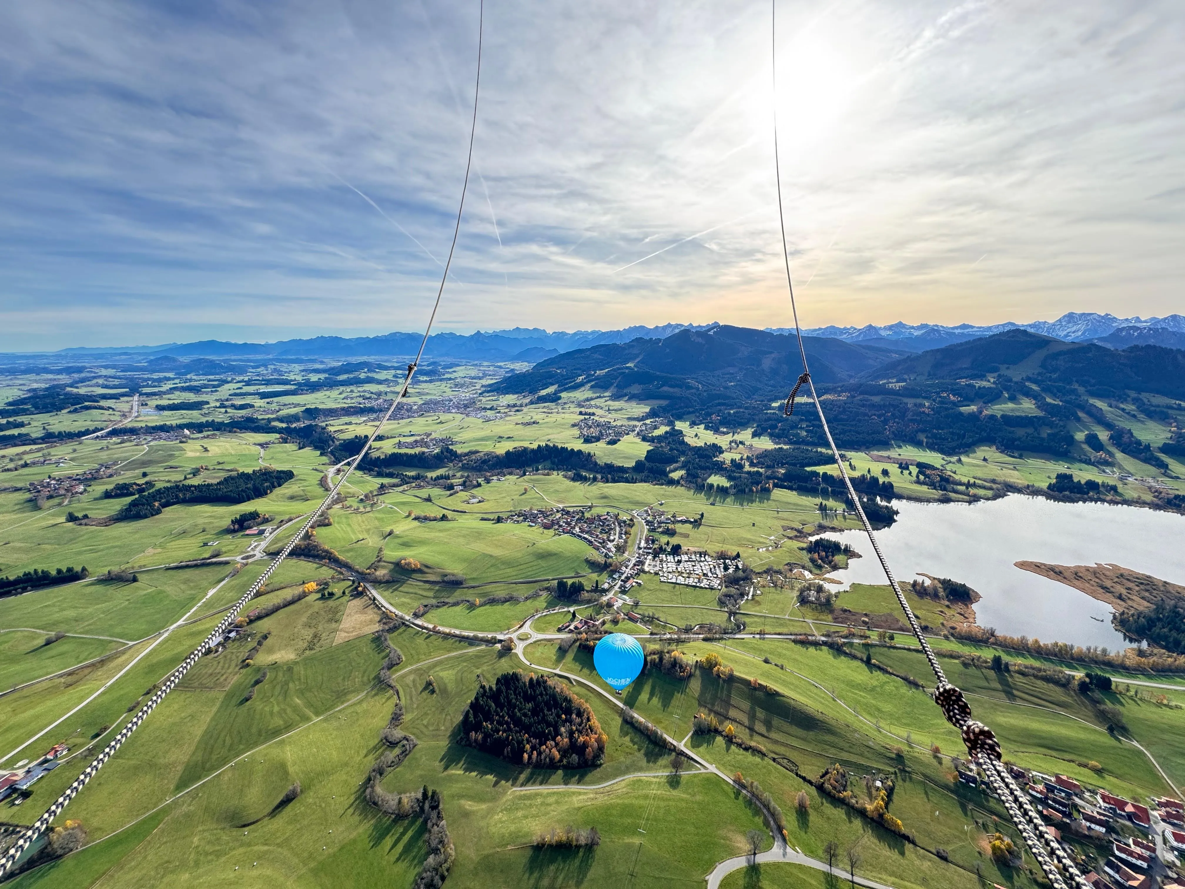 Luftaufnahme einer grünen Landschaft mit Feldern, einem See, Bergen im Hintergrund und einem blauen Heißluftballon.