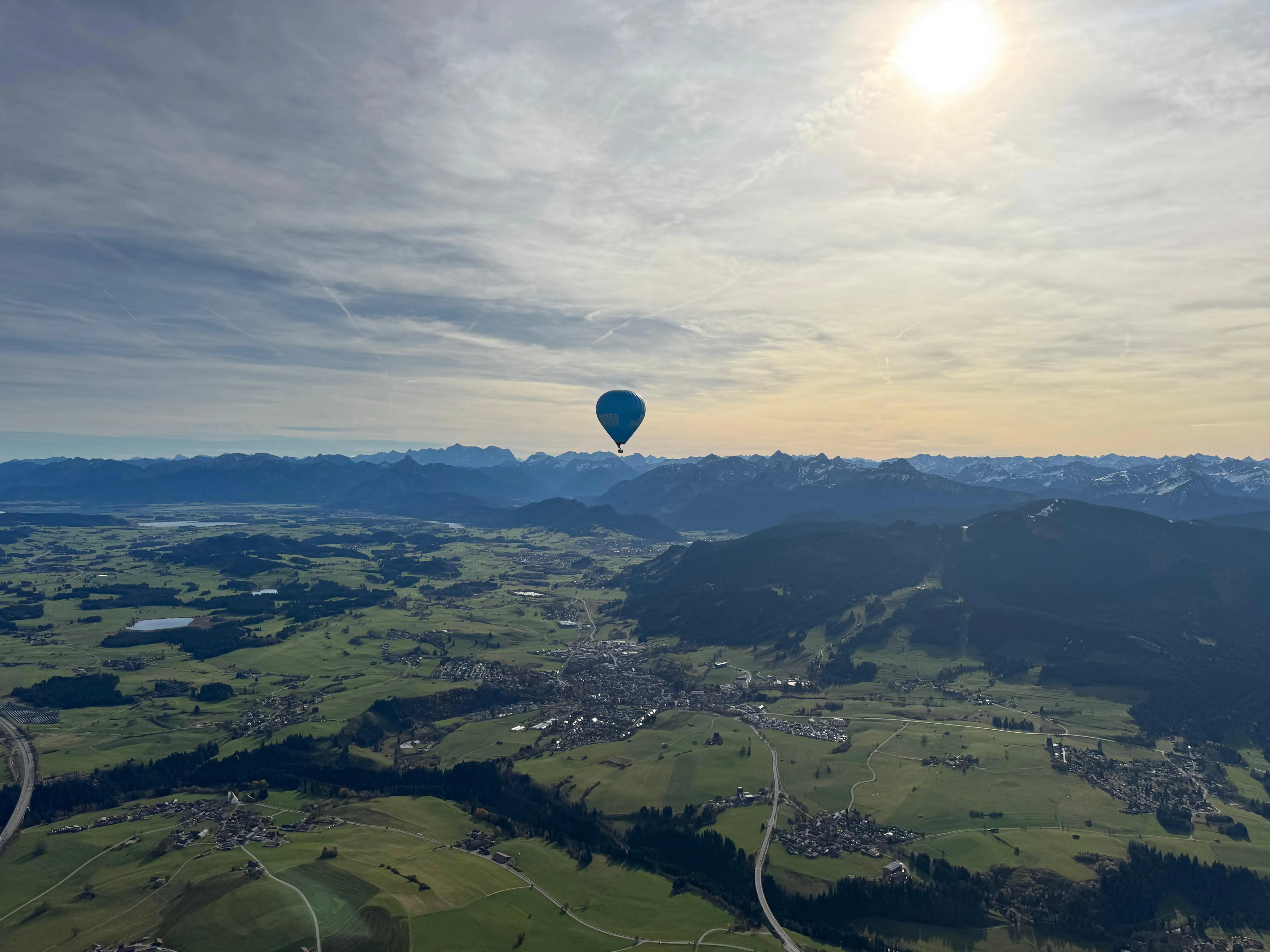 Blauer Heißluftballon schwebt über grüne Landschaft mit Straßen, Dörfern und Bergen im Hintergrund bei sonnigem Himmel.
