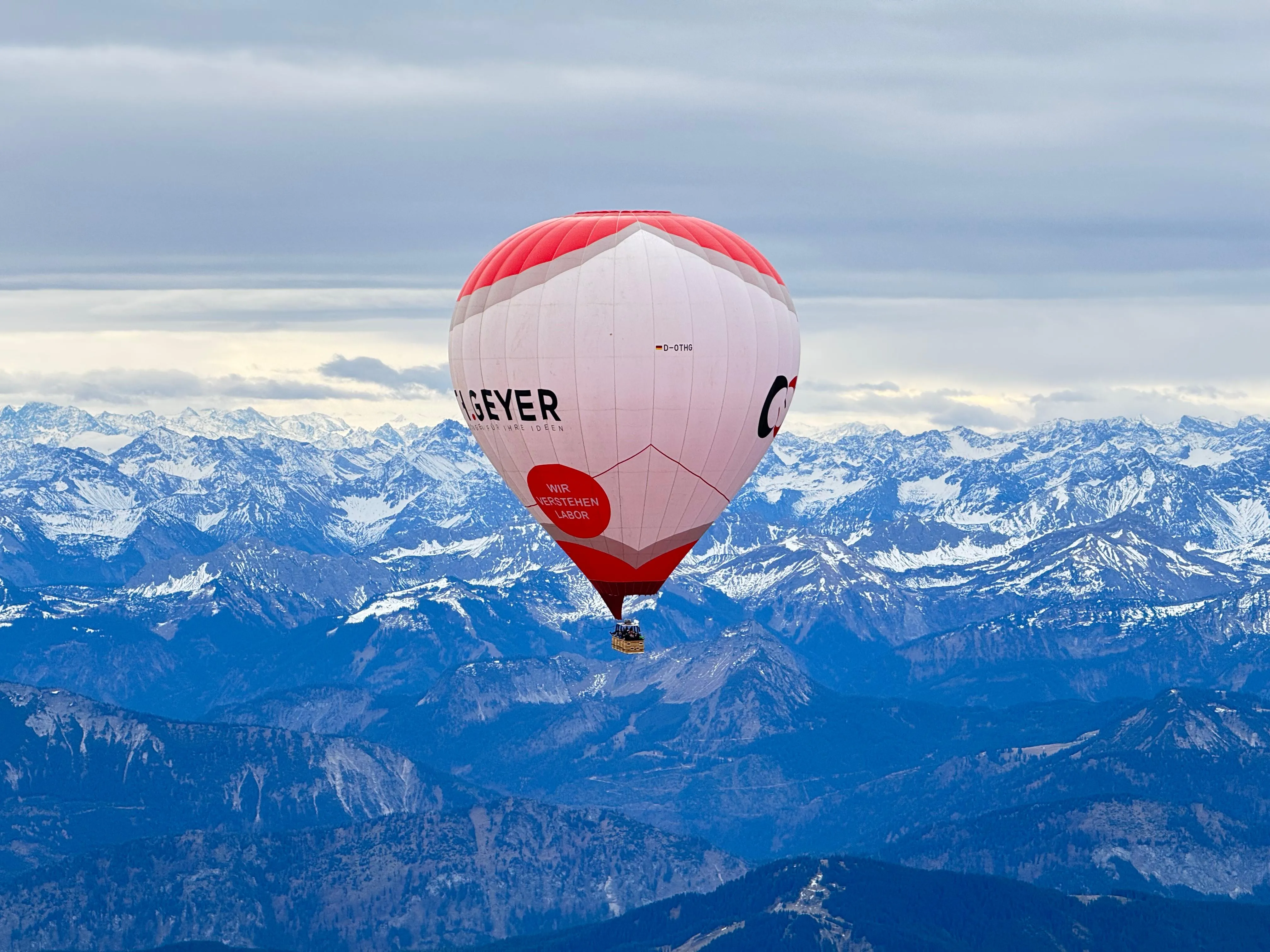 Heißluftballon mit weiß-rotem Muster fliegt über verschneite Berglandschaft unter bewölktem Himmel.