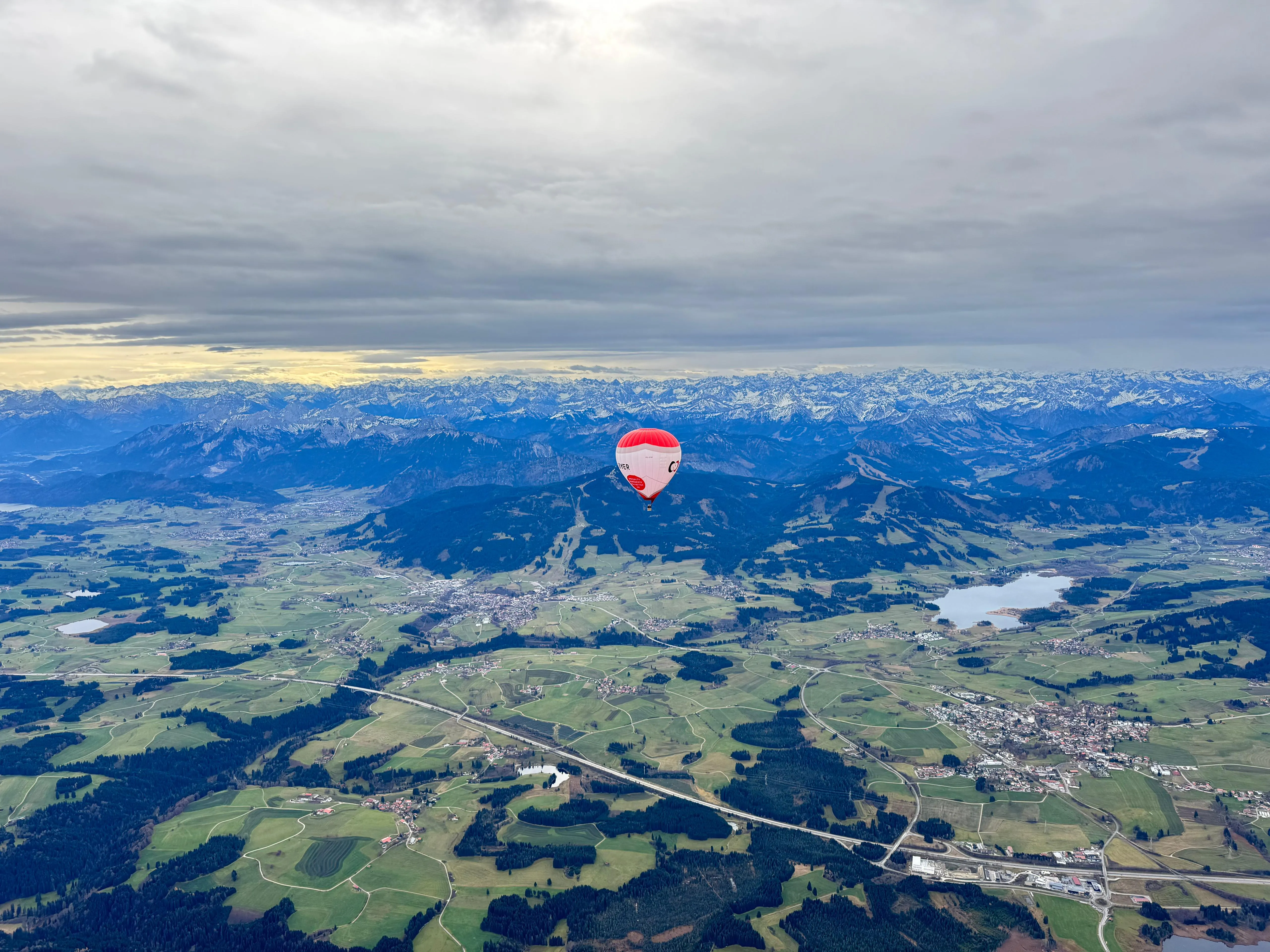 Ein roter und weißer Heißluftballon über grünen Feldern und Dörfern mit schneebedeckten Bergen im Hintergrund unter bewölktem Himmel.