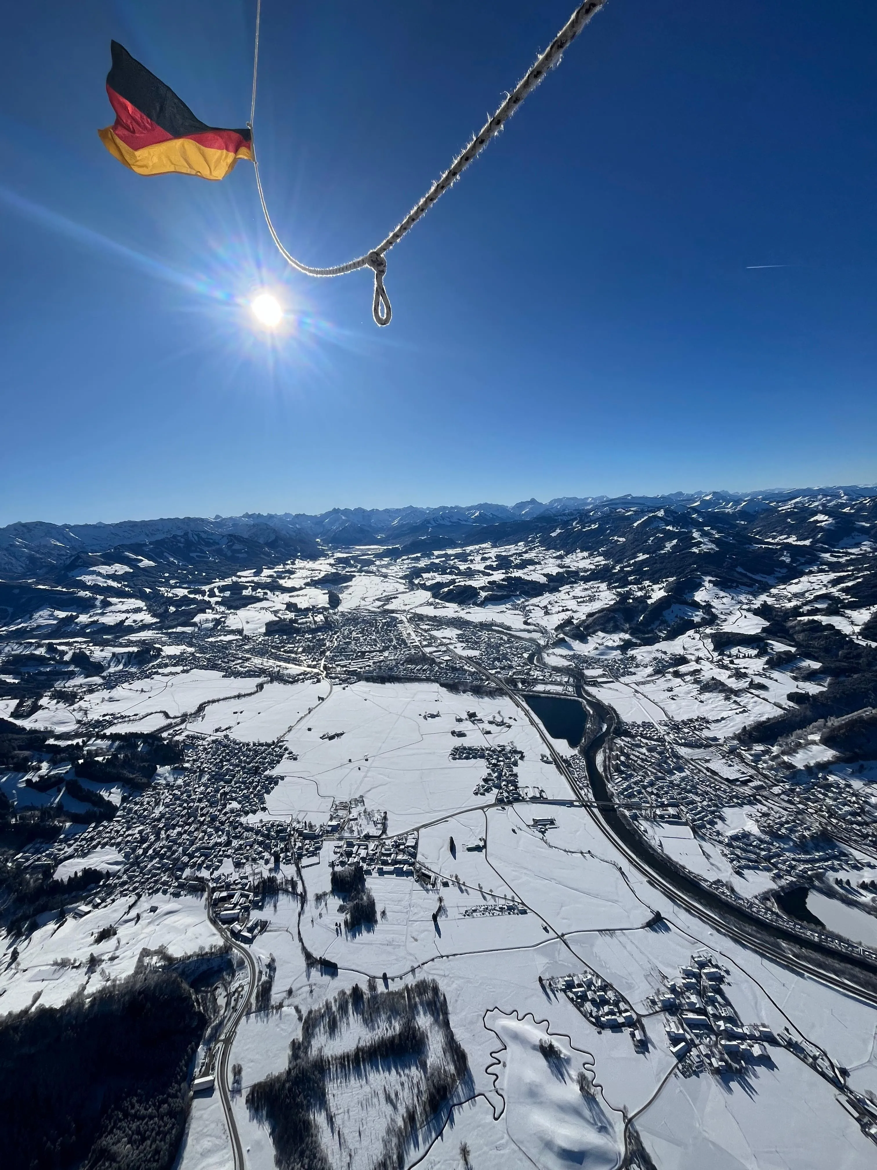 Luftaufnahme einer schneebedeckten Landschaft mit Tal, Fluss und deutschen Flagge im Vordergrund vor sonnigem blauem Himmel.