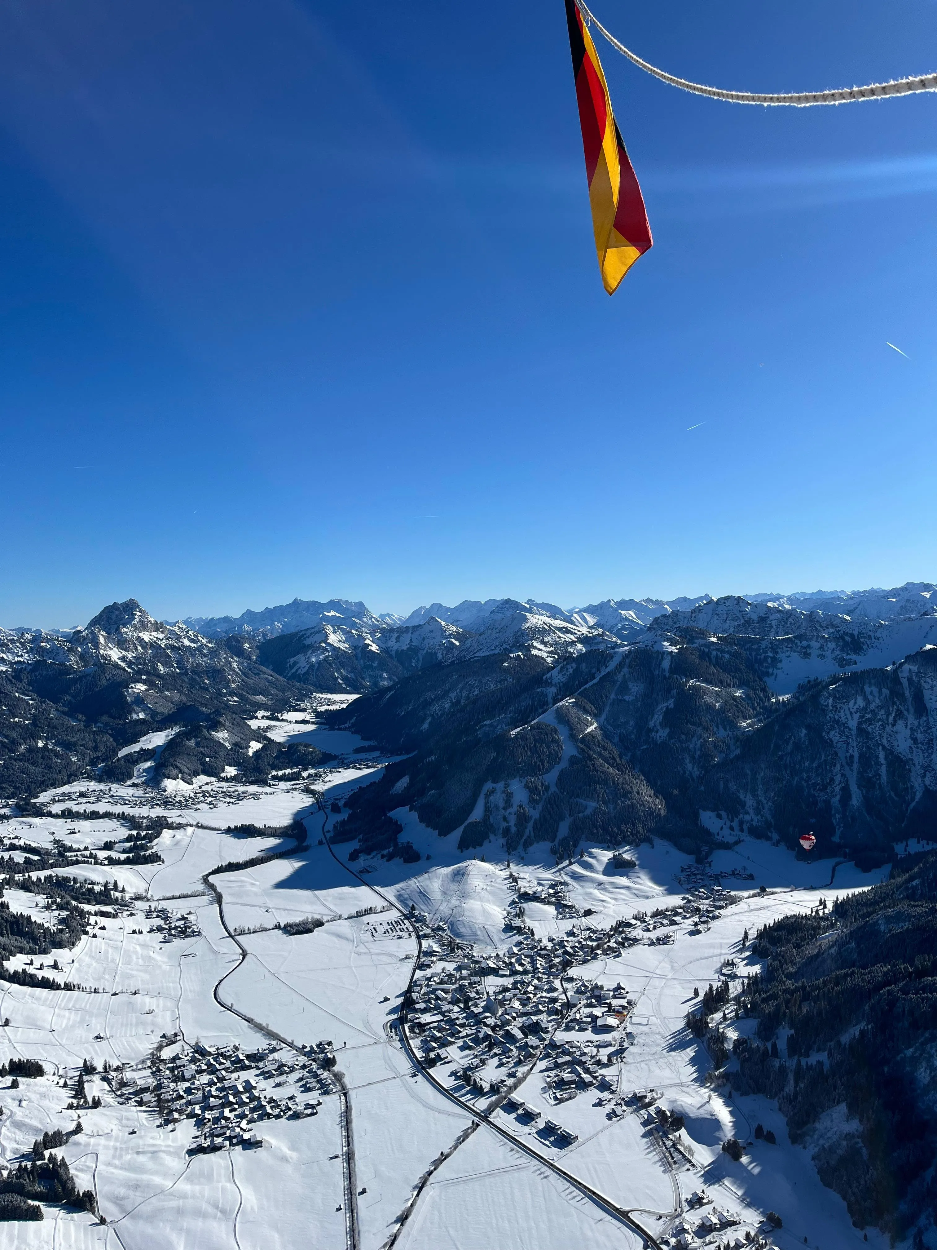 Luftaufnahme einer schneebedeckten Berglandschaft mit einem Dorf und einer deutschen Flagge oben rechts.