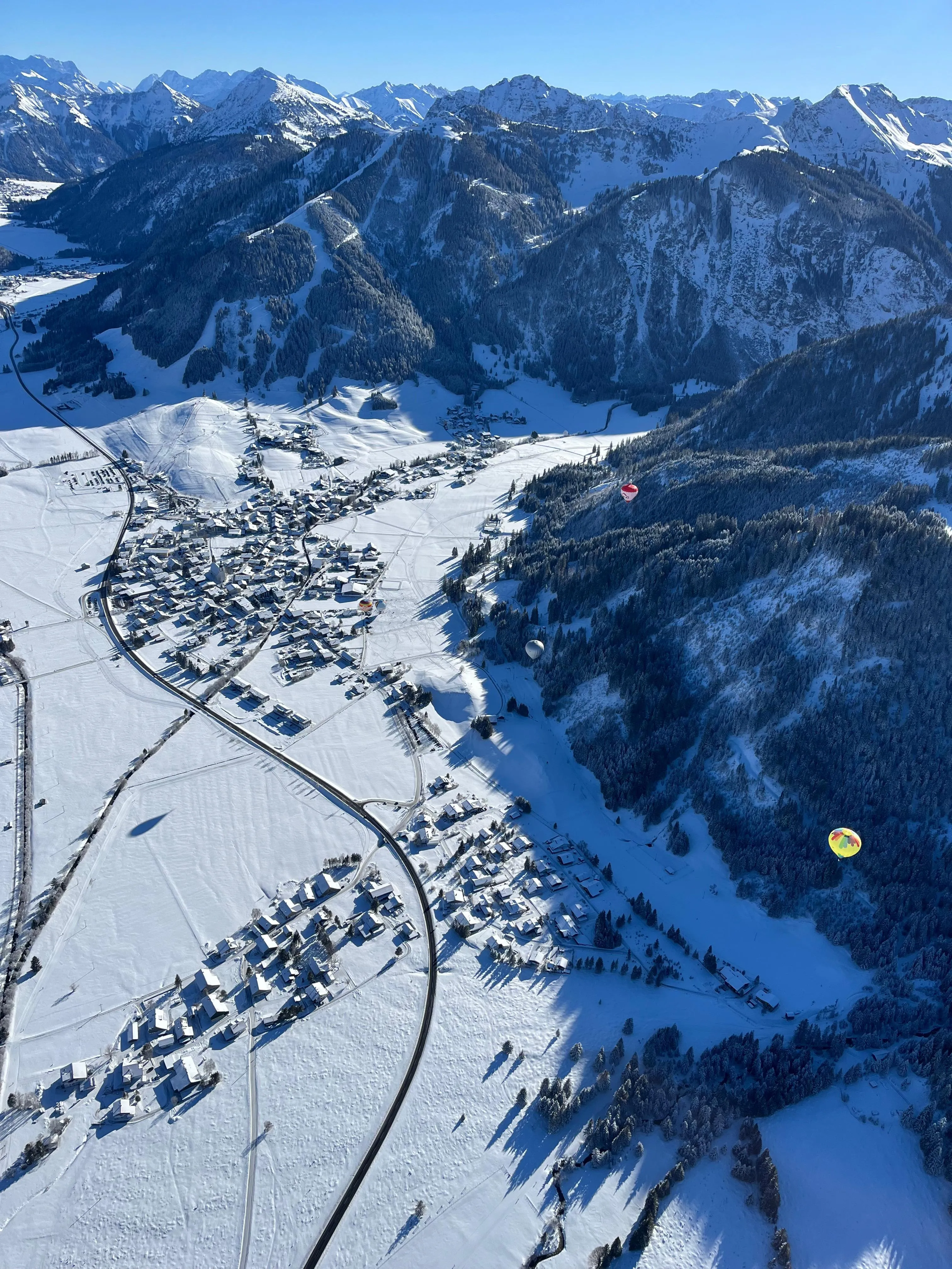 Luftaufnahme eines verschneiten Bergdorfes mit mehreren bunten Heißluftballons über verschneiten Wäldern und Bergen.