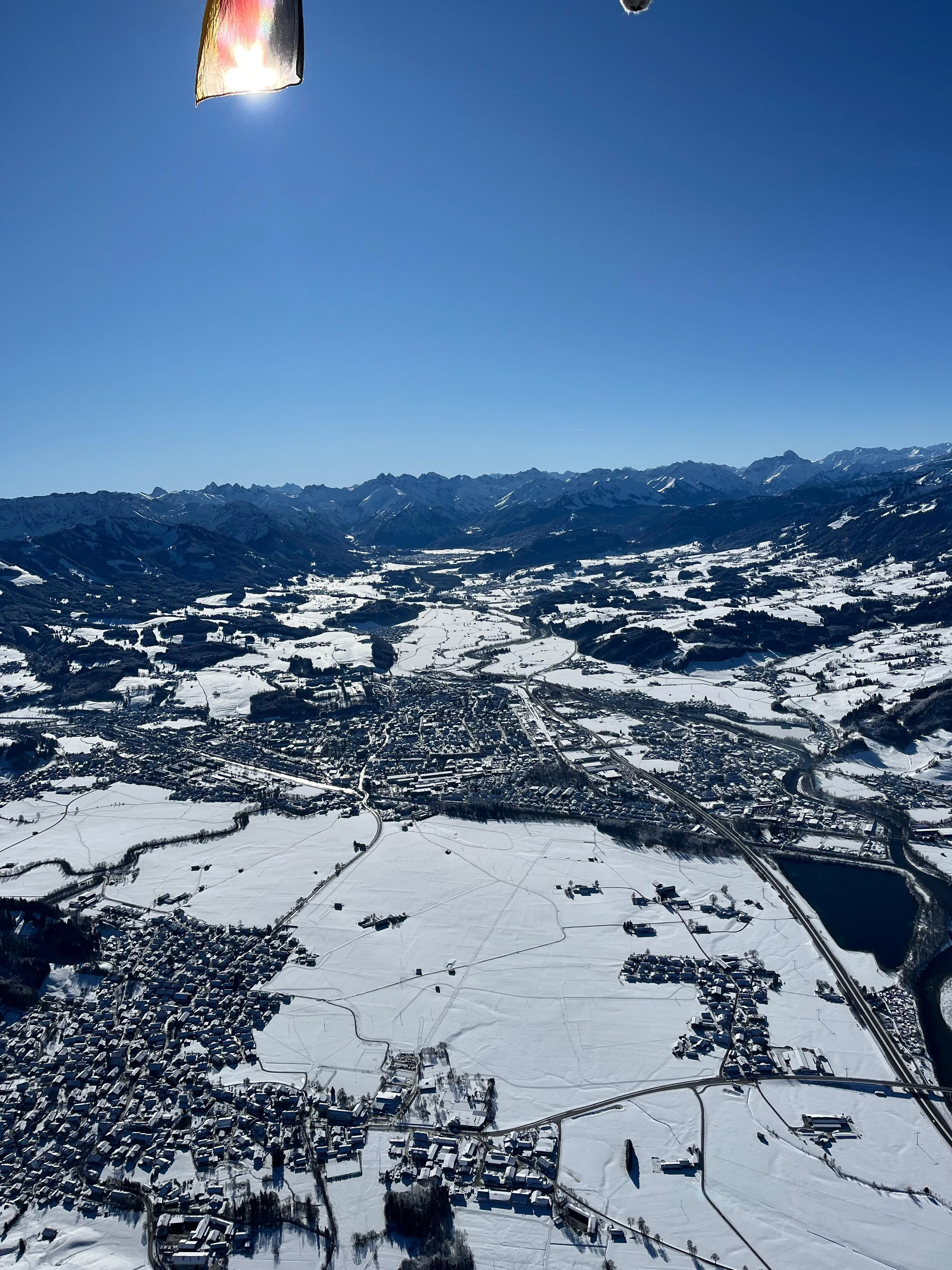 Vogelperspektive auf schneebedeckte Stadt und Felder mit Bergen im Hintergrund unter klarem blauem Himmel.