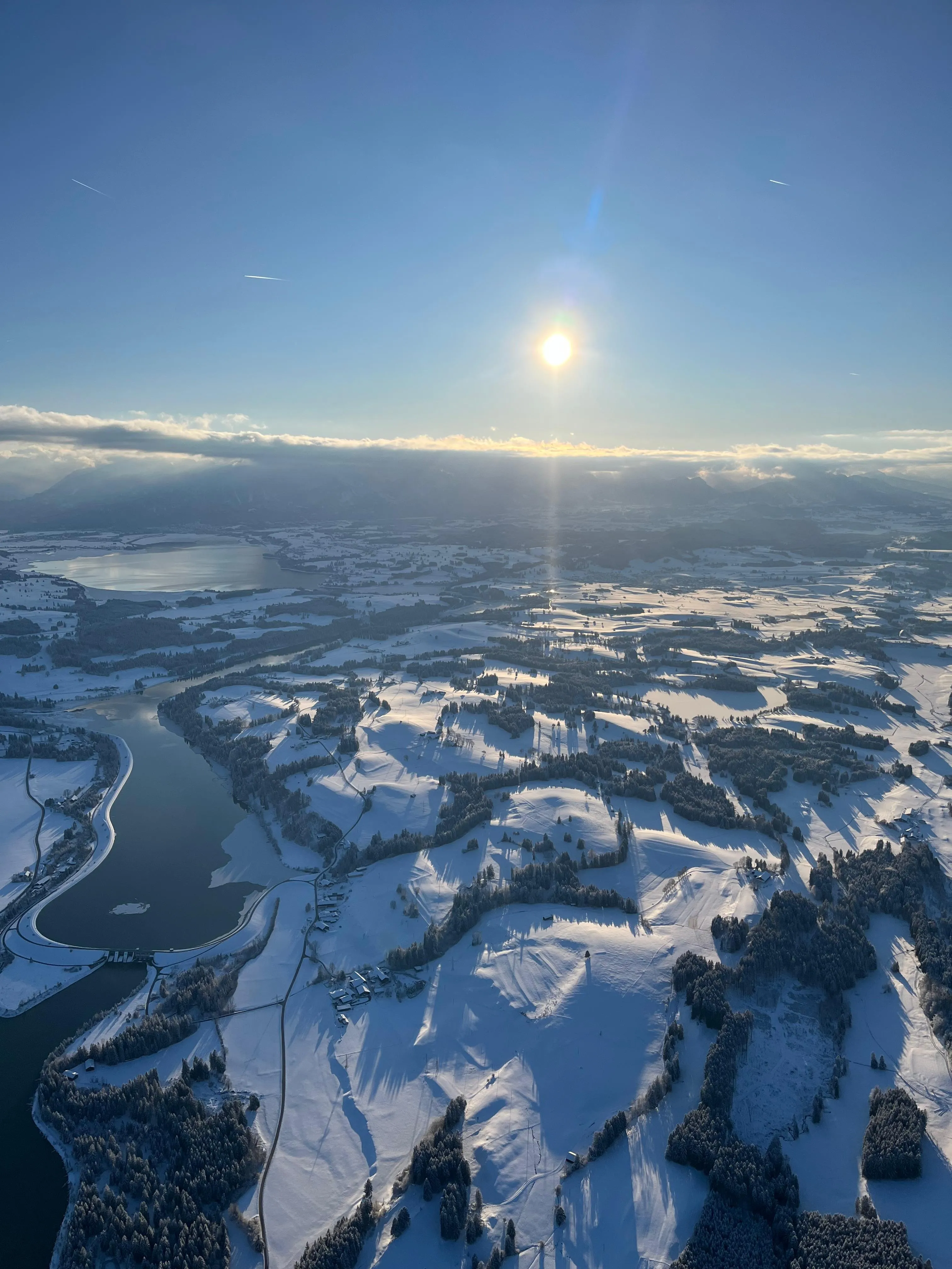Luftaufnahme einer verschneiten Landschaft mit Seen, Feldern und Wäldern bei klarem Himmel und tiefstehender Sonne.
