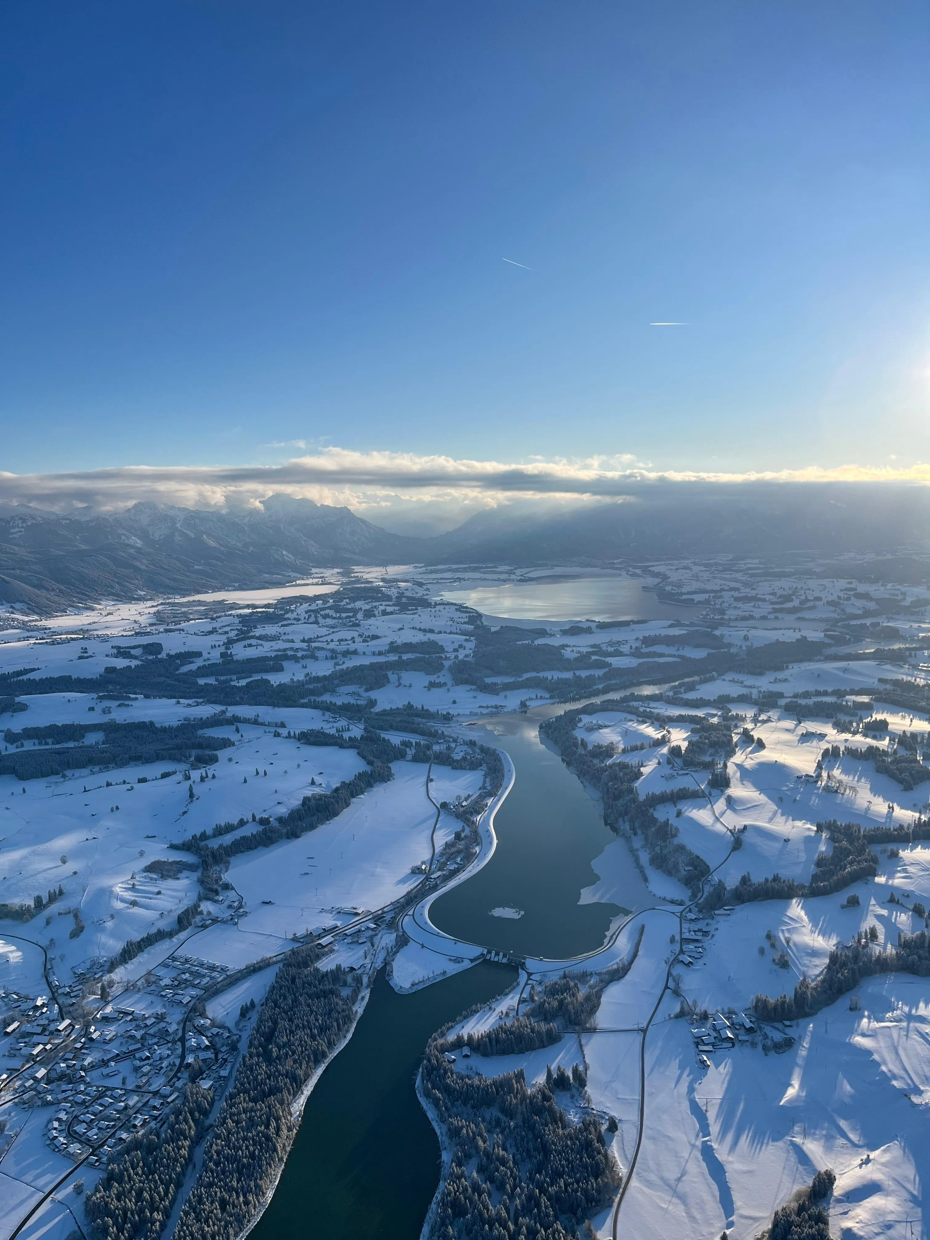 Luftaufnahme einer verschneiten Landschaft mit Fluss, Seen, Wäldern und Bergen unter blauem Himmel bei Sonnenaufgang.