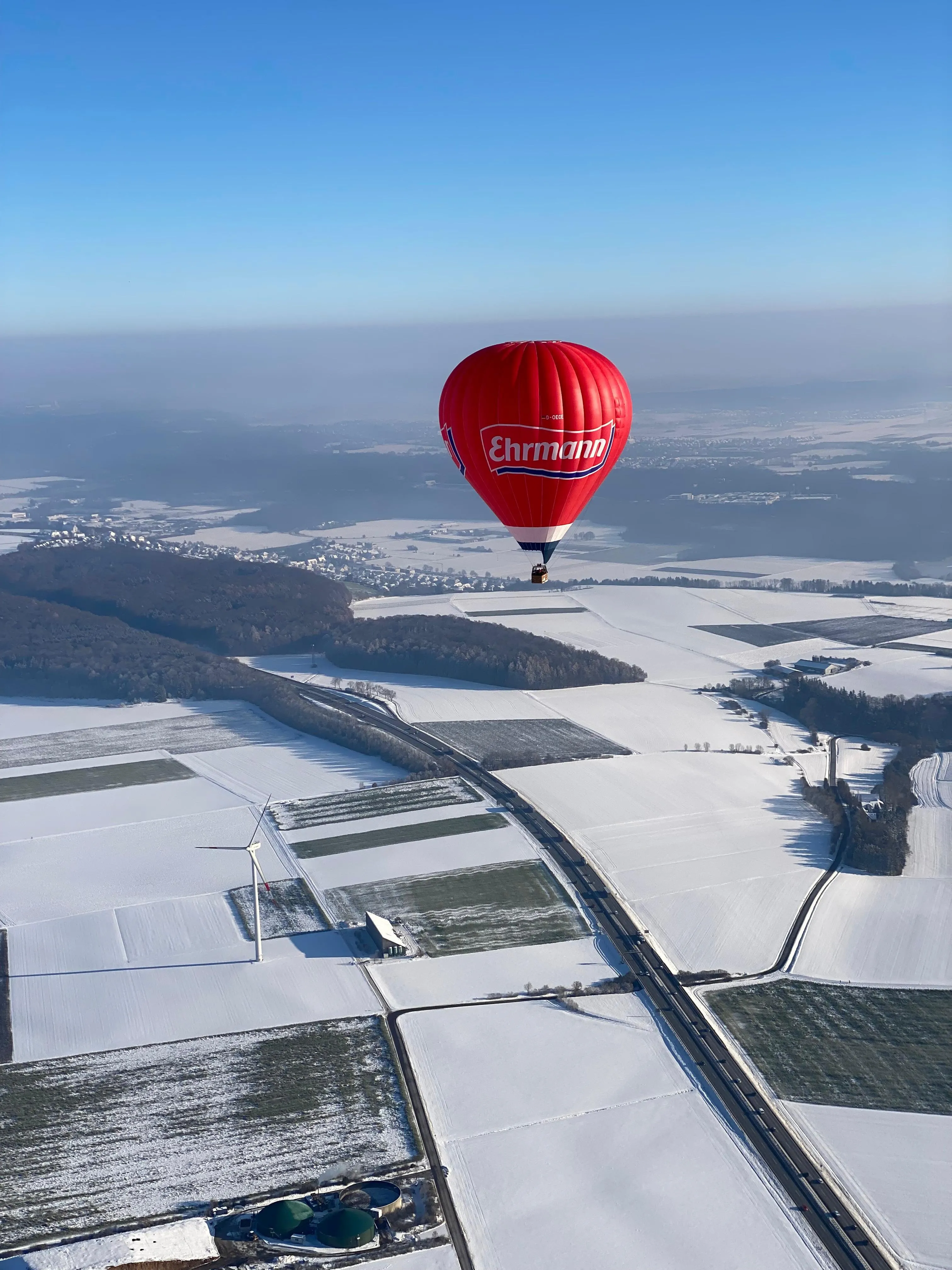 Roter Heißluftballon mit dem Schriftzug Ehrmann fliegt über schneebedeckte Felder und eine Autobahn unter blauem Himmel.