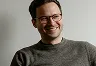 Lächelnder Mann mit Brille trägt graues Sweatshirt vor hellem Hintergrund.