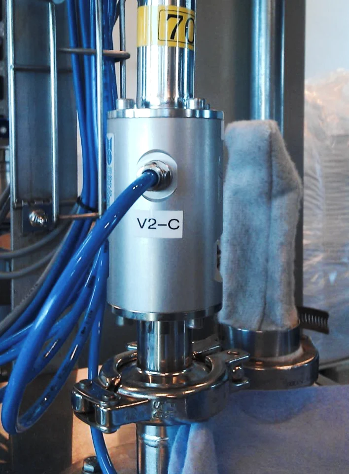 Industrielles Metallventil mit der Beschriftung V2-C, verbunden mit blauen Kabeln und einer abgedeckten Rohrleitung.