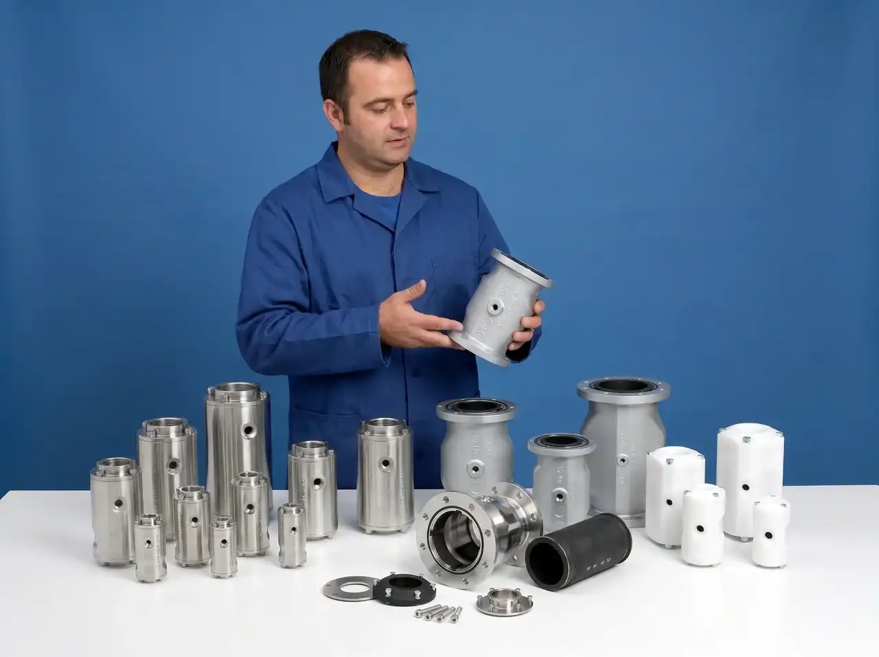 Mann in blauem Overall hält großes Metallventil, mehrere Metall- und Kunststoffventile auf Tisch.