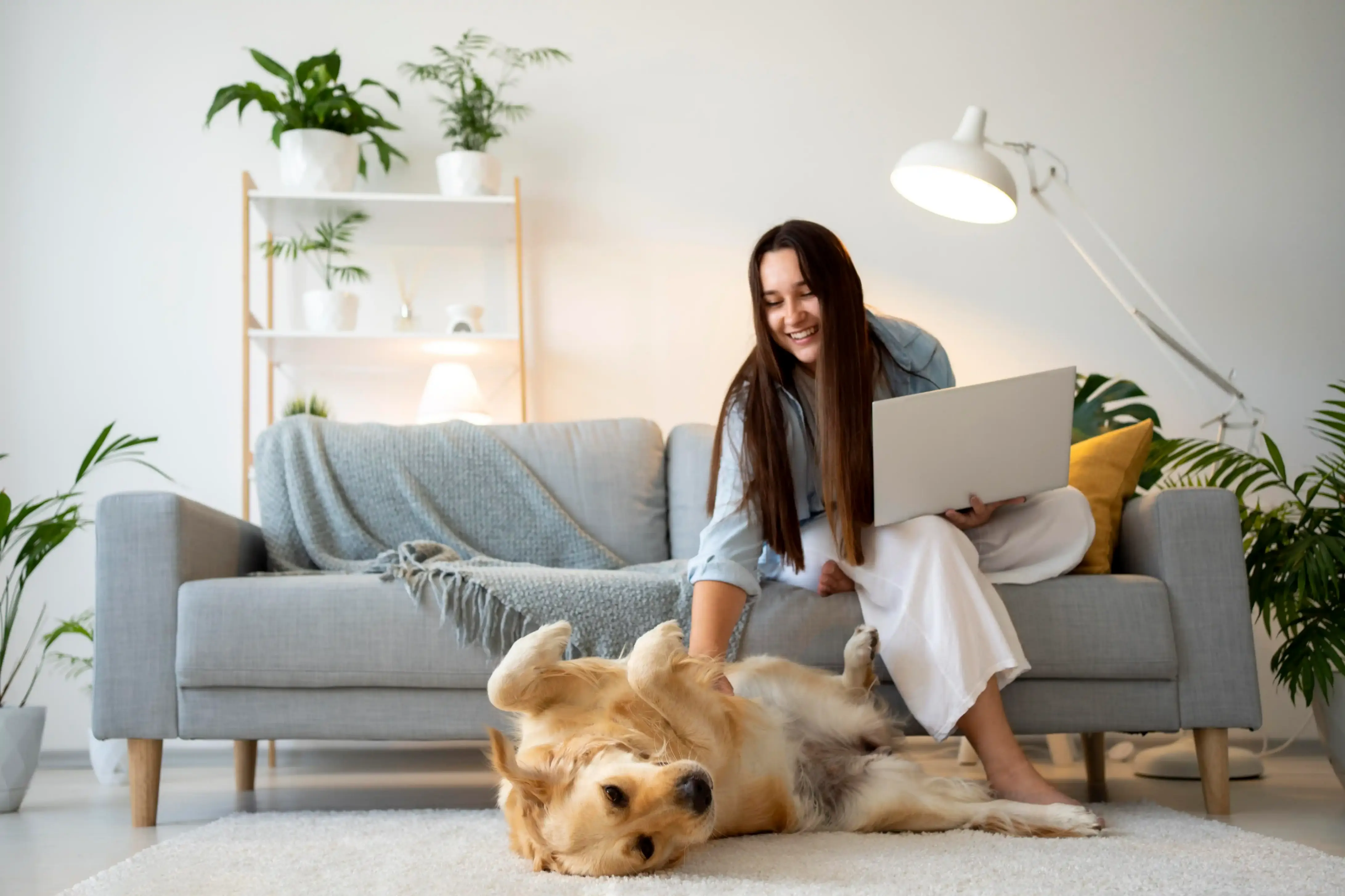 Une jeune femme assise sur le canapé essaie de travailler sur l'ordinateur, elle caresse le chien allongé à ses pieds.