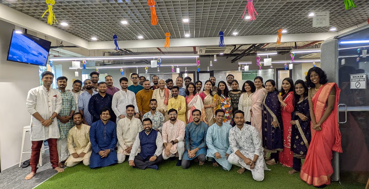 Group photo of men and women in colorful traditional Indian clothing standing and sitting indoors on artificial green grass under a ceiling decorated with hanging tassels.