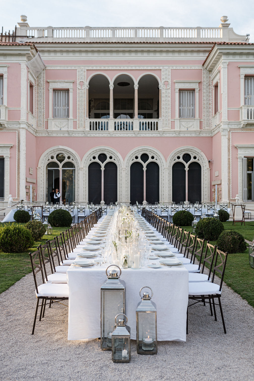 Grande table blanche dressée pour un dîner en extérieur devant une maison de style classique rose avec de grandes fenêtres en arche.