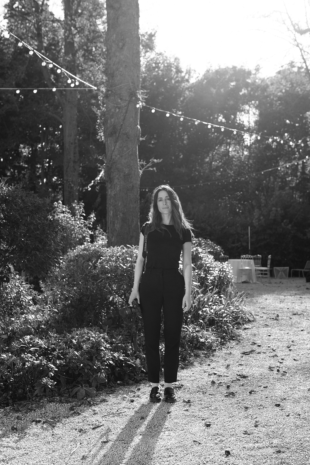 Woman standing on a gravel path in a garden holding a camera, with string lights overhead and trees in the background.
