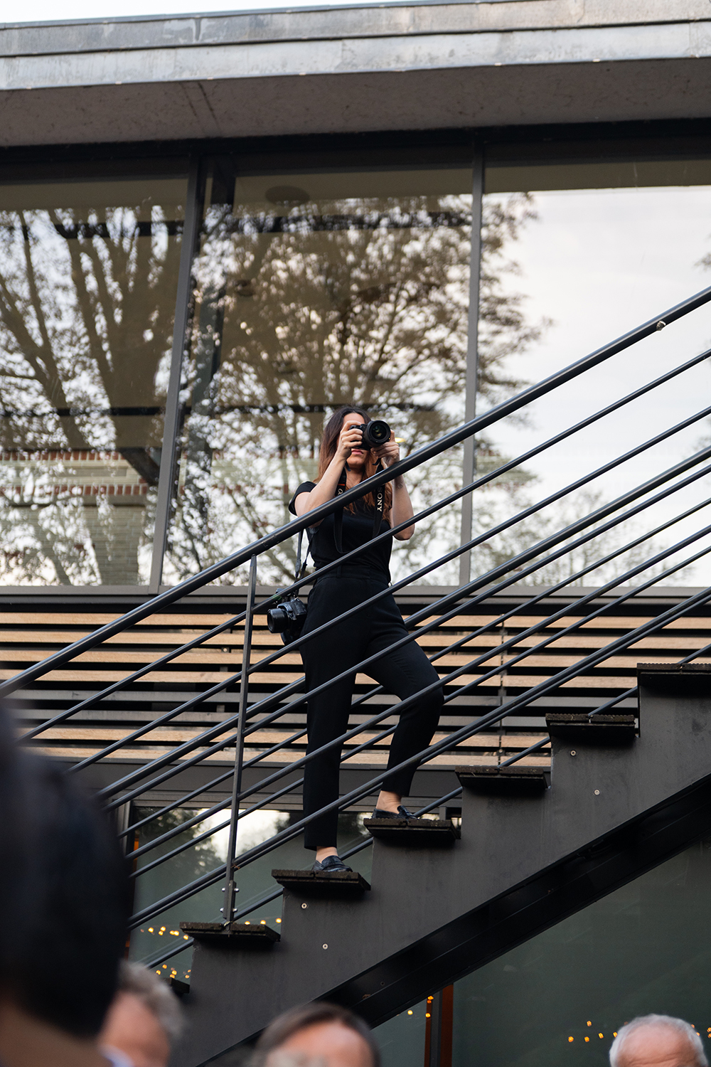 Portrait of Marie-Louise Photographer based in Paris. Description of the image is Woman dressed in black standing on outdoor stairs taking a photo with a camera, with reflections of trees in the building's glass windows behind her.