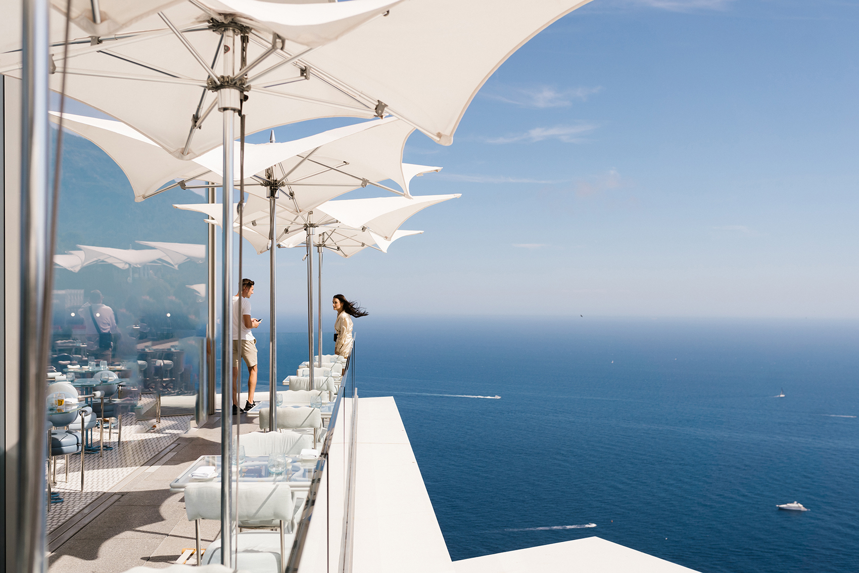 Two people standing on a balcony with white umbrellas overlooking a vast blue sea with boats in the distance.