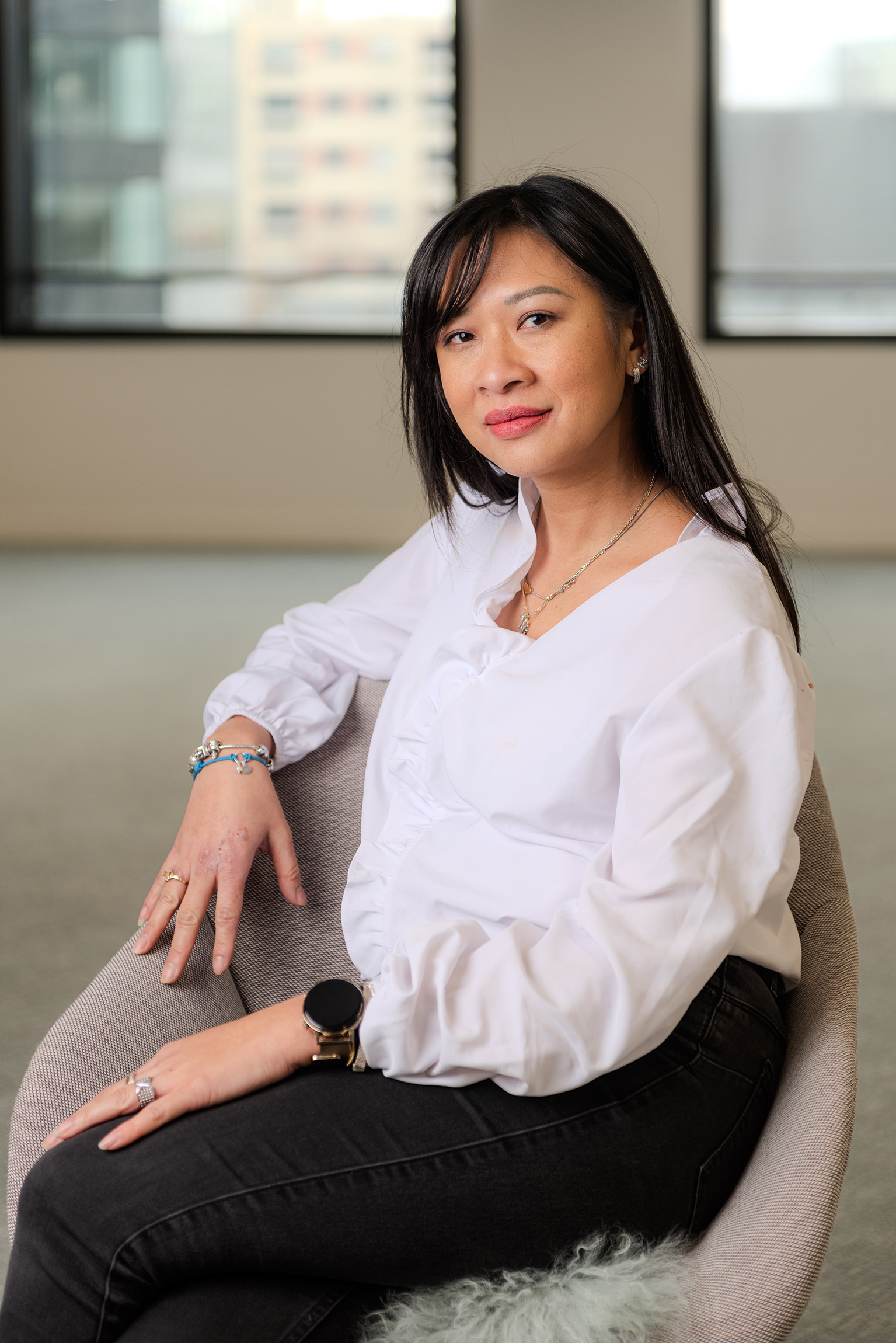 Woman with long dark hair wearing a white blouse and black jeans sitting comfortably in a beige chair indoors.