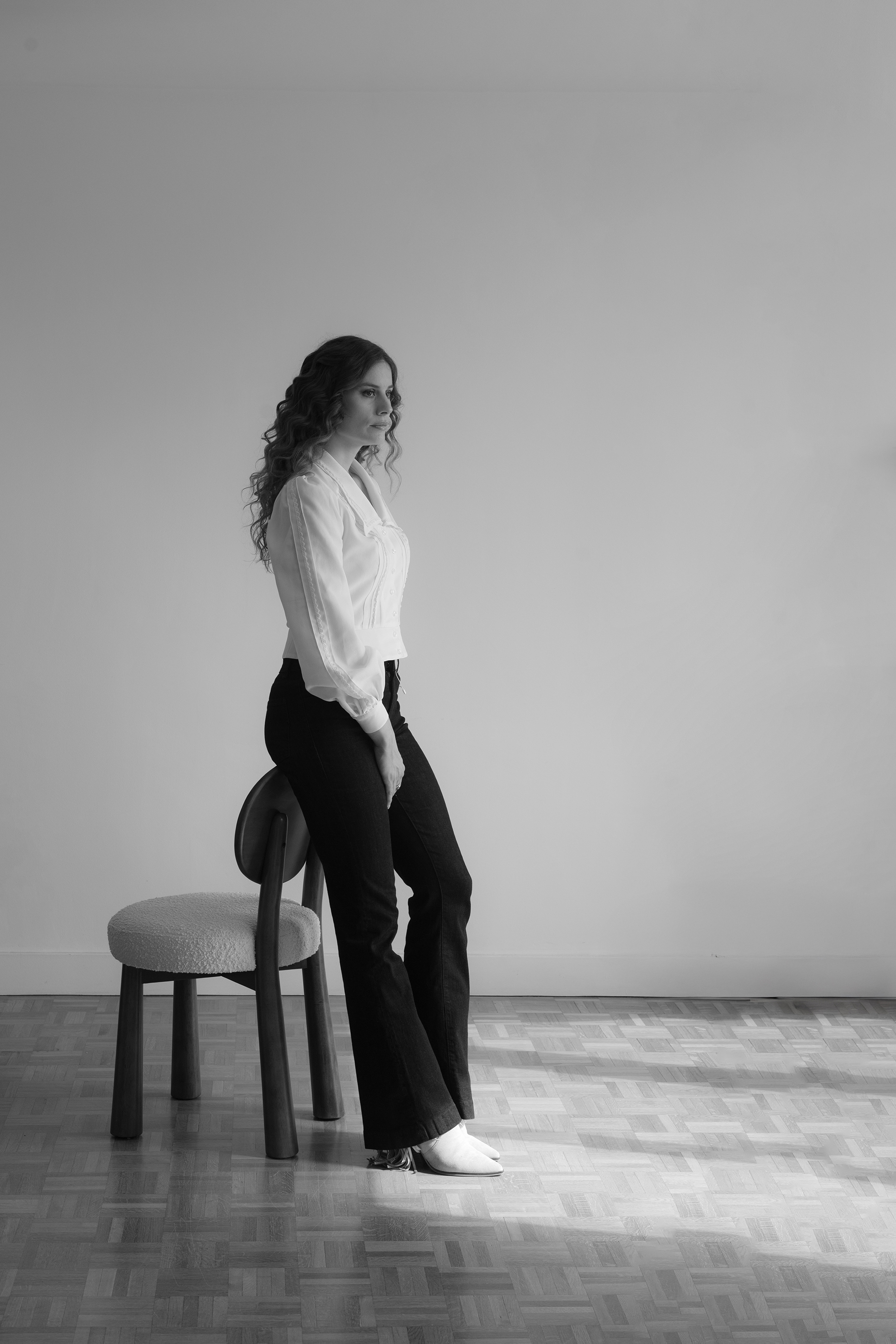 Femme assise de profil sur une chaise, portant une blouse blanche et un pantalon noir, regardant devant elle dans une pièce claire.