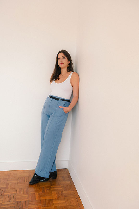 Smiling woman with long wavy brown hair resting her face on her hand against a plain light background.