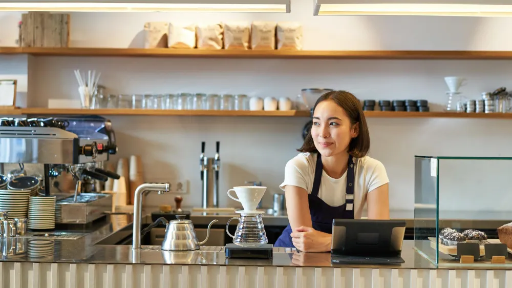 Barista i modern kaffebar som förbereder kaffe bakom disken med espressomaskin och bakverk i glasmonter.