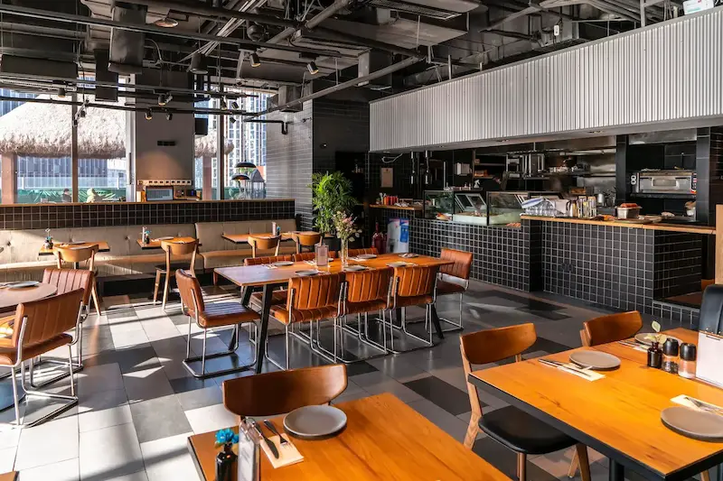 Ljus och modern restaurang med öppet kök, träbord, bruna läderstolar och stilren industriell inredning i soligt dagsljus.