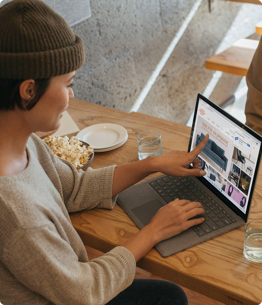 Persona sentada en una mesa de madera, usando una laptop y señalando con el dedo una página web de muebles en la pantalla.