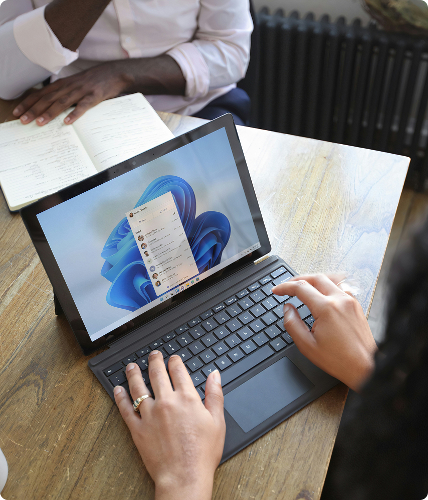 Una persona usando una laptop con Windows 11 sobre una mesa de madera mientras otra toma notas en un cuaderno.