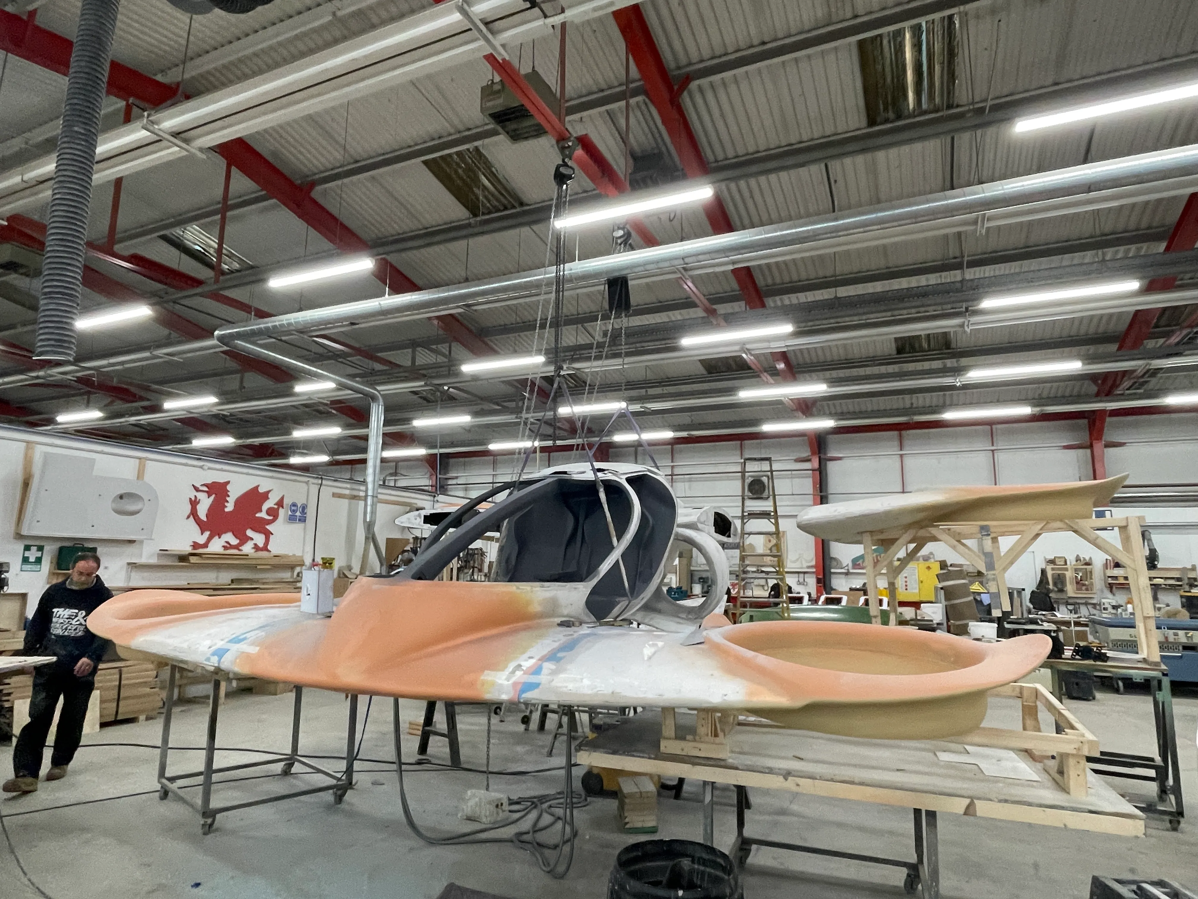 Early-stage assembly of Doroni’s aircraft prototype inside a workshop, with cockpit structure suspended from a ceiling hoist and wings under construction nearby.
