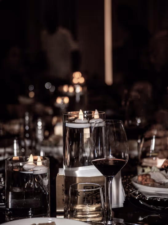 Table setting with floating candles in glass holders, a glass of red wine, and dim ambient lighting in the background.